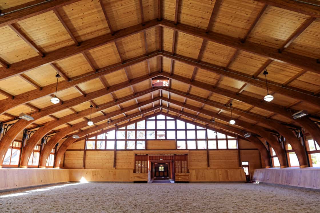 A spacious luxury horse barn featuring high, arched wooden beams, large windows that flood the indoor riding arena with natural light, and sand-covered flooring. The entryway is visible at the far end.