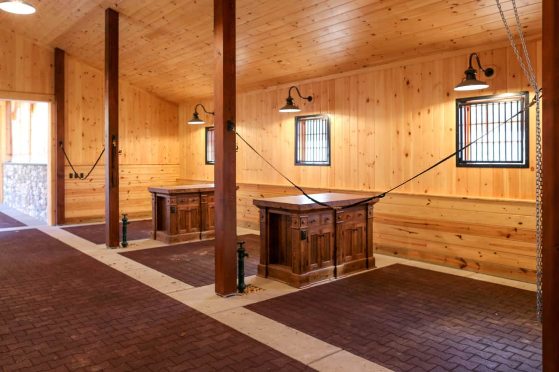 A clean, modern luxury horse barn with wooden walls, brick flooring, two wooden grooming stations, overhead lamps, and windows with metal bars letting in natural light.
