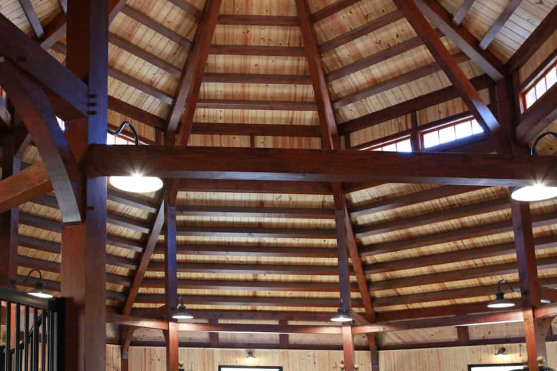 Interior view of a round, wooden-roofed luxury horse barn with black metal gates and wooden walls, featuring a high, vaulted ceiling with exposed beams and natural light streaming through upper windows.
