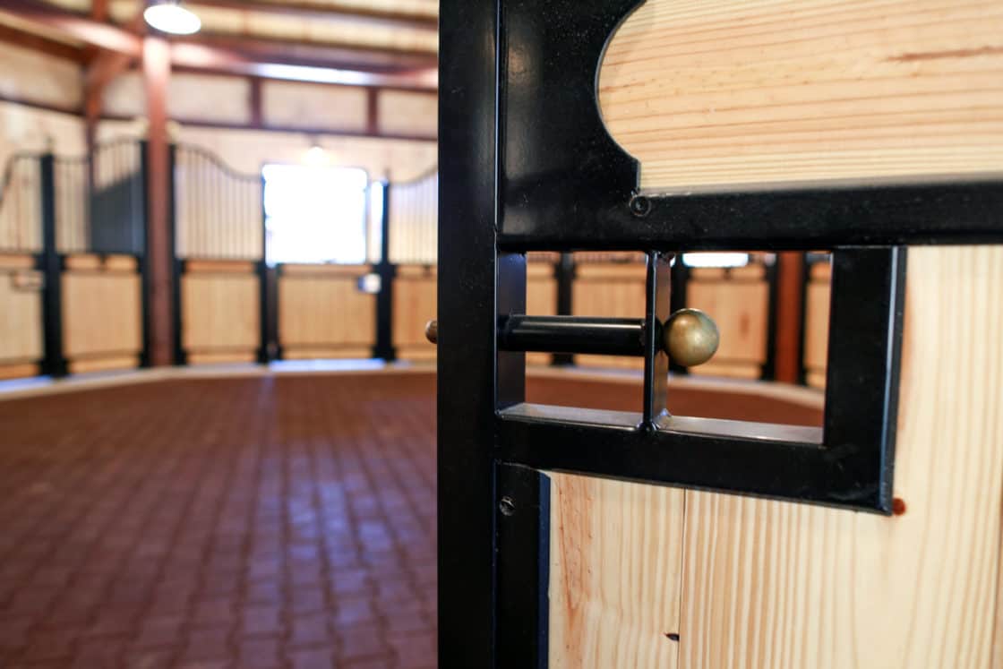Close-up of a metal and wood latch on a stall door inside a clean, well-lit luxury horse barn with brick flooring and other stalls visible in the background.
