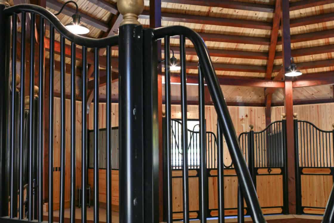 Interior of a luxury horse barn with tall ceilings, exposed beams, and elegant black metal and wood stall doors under bright overhead lights.