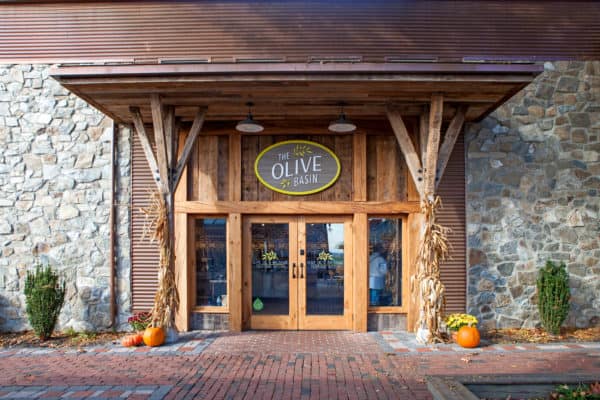 The entrance of The Olive Basin store features double wooden doors and stone and wood walls reminiscent of a luxury horse barn, with autumn decorations like cornstalks, pumpkins, potted shrubs, and a large oval sign above the door.