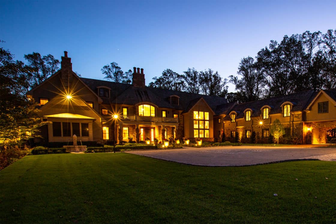 Large, elegant mansion with glowing lights at dusk, featuring multiple windows and chimneys, plus an expansive driveway bordered by grass and trees—perfectly complemented by a stylish luxury horse barn nearby.
