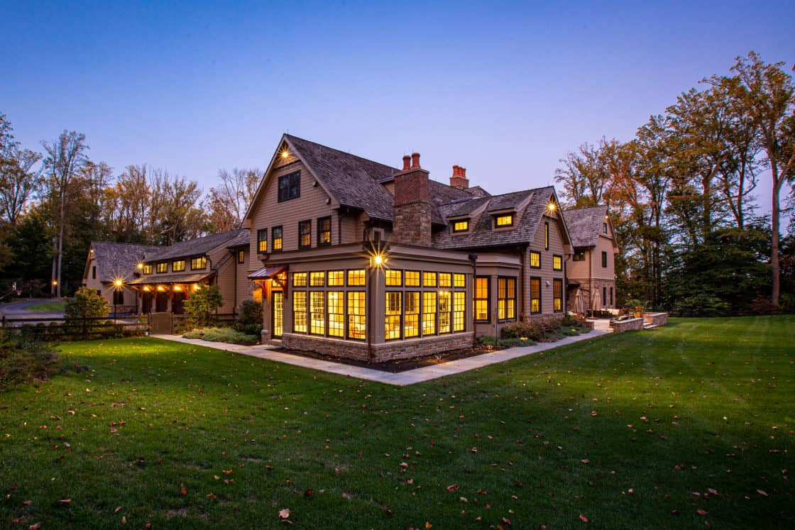 Large, multi-story luxury home with warm yellow lights glowing from numerous windows at dusk, surrounded by green lawn and tall trees, under a clear evening sky.