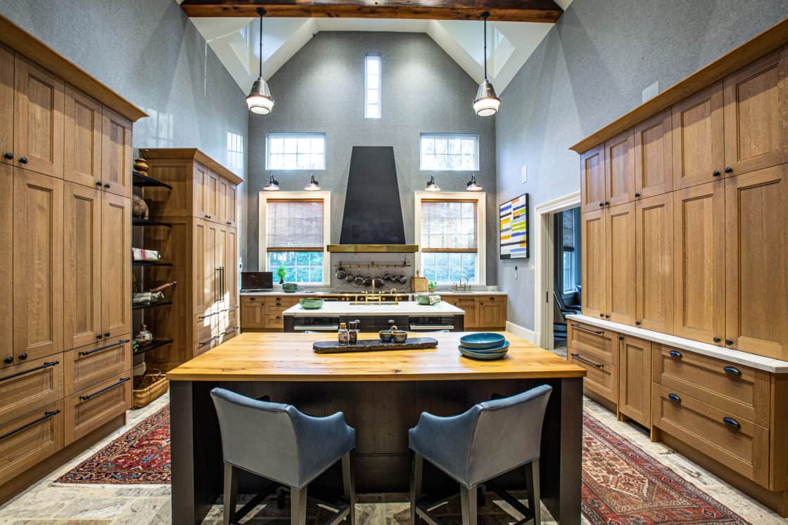 Modern kitchen with high ceilings, wooden cabinets, a large island and blue chairs, sleek stainless steel appliances, a black range hood, and decorative rugs. Natural light pours in—perfect for entertaining in this luxury home or modern party barn.