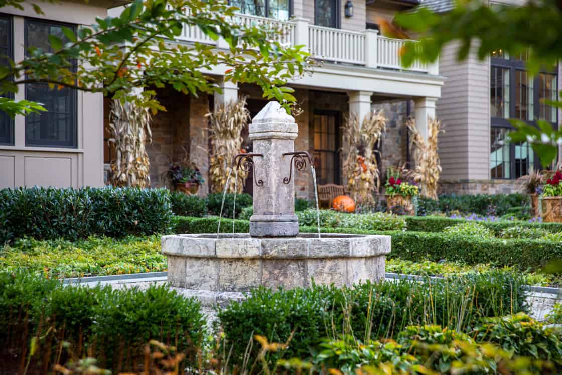 A stone fountain with water streams stands in a manicured garden, surrounded by trimmed bushes. In the background is a luxury home with a porch, autumn corn stalk decorations, and potted plants.