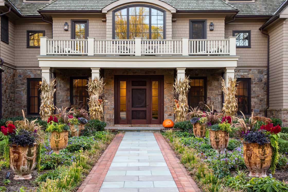 A stone and shingle house with a front porch, decorated for autumn with cornstalks, potted plants, and a pumpkin by the door. A brick-edged walkway leads through a lush garden to the entrance, beside a charming bank barn.