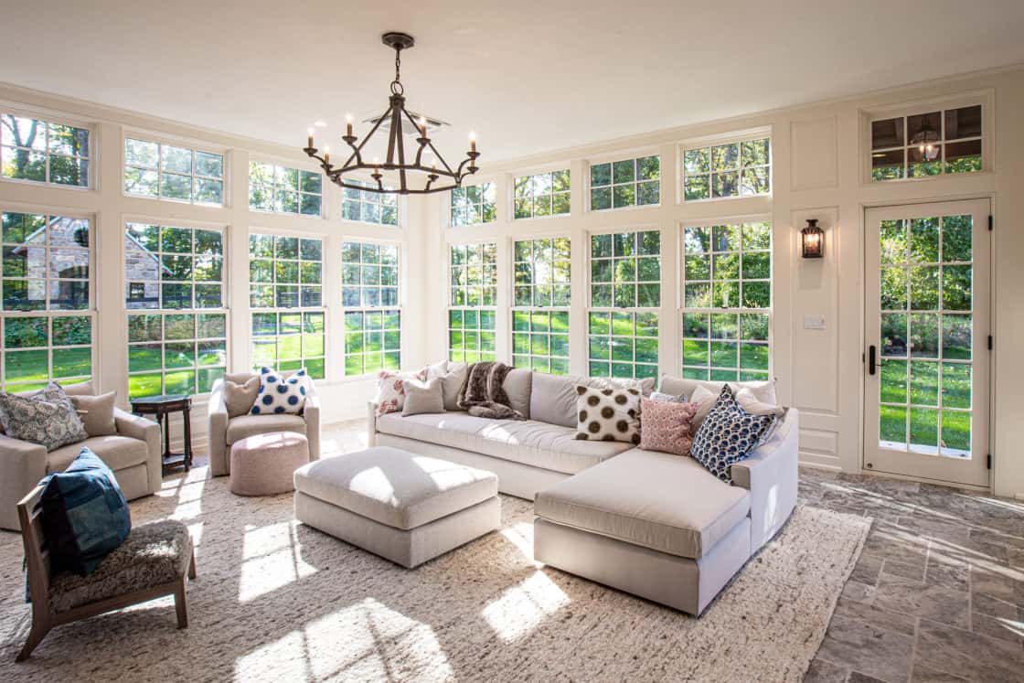Bright sunroom with large windows in a luxury home, featuring a white sectional sofa with patterned pillows, a light ottoman, cozy armchairs, and a rug. Green lawn and trees are visible outside. A chandelier hangs from the ceiling.