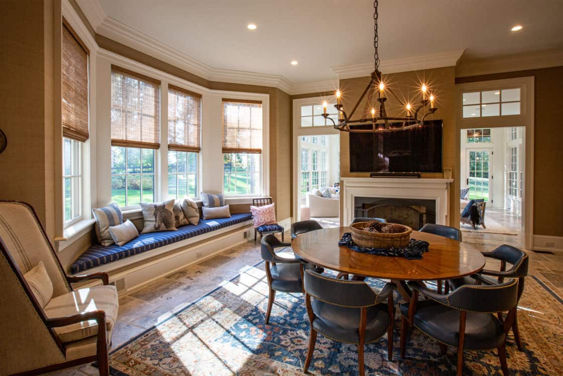 A cozy, sunlit living and dining area in this luxury home features a large bay window seat, round wooden dining table, six chairs, fireplace with TV above, chandelier, and views into a sitting room and garden. A dog stands in the adjacent room.