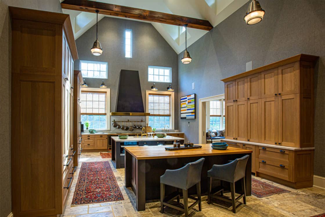 Spacious modern kitchen with high vaulted ceiling, wood cabinets, large island with two blue chairs—reminiscent of a luxury horse barn—pendant lights, and multiple windows letting in natural light. Oriental rugs line the tiled floor.