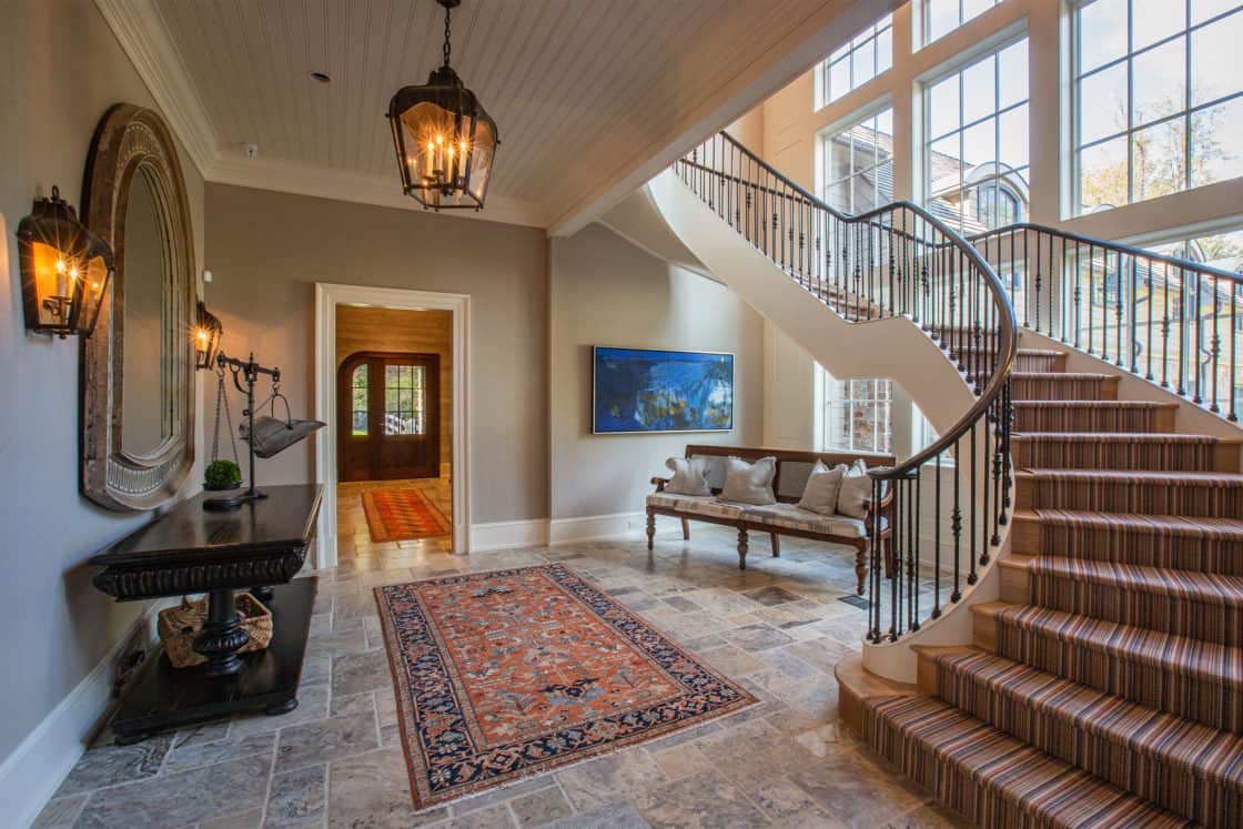 Elegant entryway with a patterned rug on stone flooring, a curved staircase with striped carpet, and large windows reflects the charm of a luxury home—complete with lantern lights and rustic touches inspired by a classic bank barn.