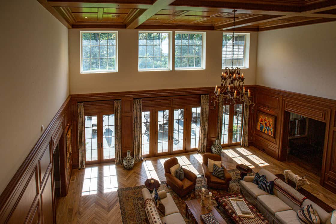 Spacious living room with high ceilings, large upper windows, wooden paneling, and floor-to-ceiling glass doors. This luxury home features cozy seating, a chandelier, rugs, decorative vases, and wall art. Sunlight streams in, creating bright patterns.