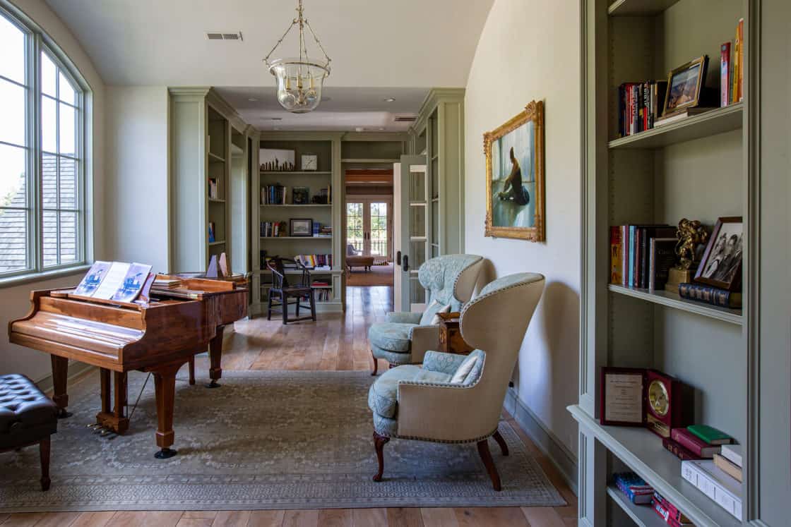 A cozy luxury home library with a grand piano, two upholstered armchairs, bookshelves brimming with books and decor, large windows, and framed artwork—an elegant, well-lit room with hardwood floors perfect for relaxing or entertaining.