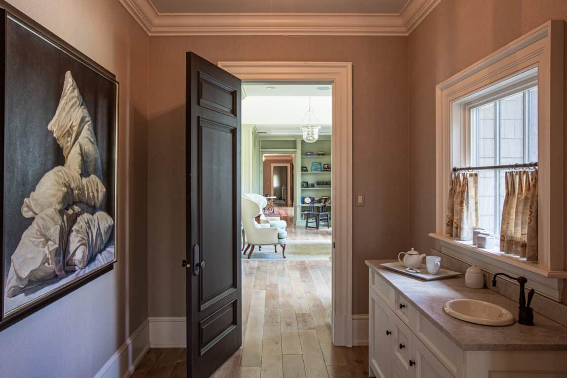 A hallway in a luxury home features a painting of stacked pillows, a white sink and tea set on the counter, and an open black door leading to a sunlit living room with an armchair and built-in shelves.