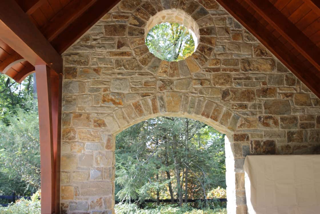 A covered stone patio with a wooden ceiling features wicker furniture, two lanterns, and a circular window above an arched opening that looks out onto the green, wooded yard of a luxury home.
