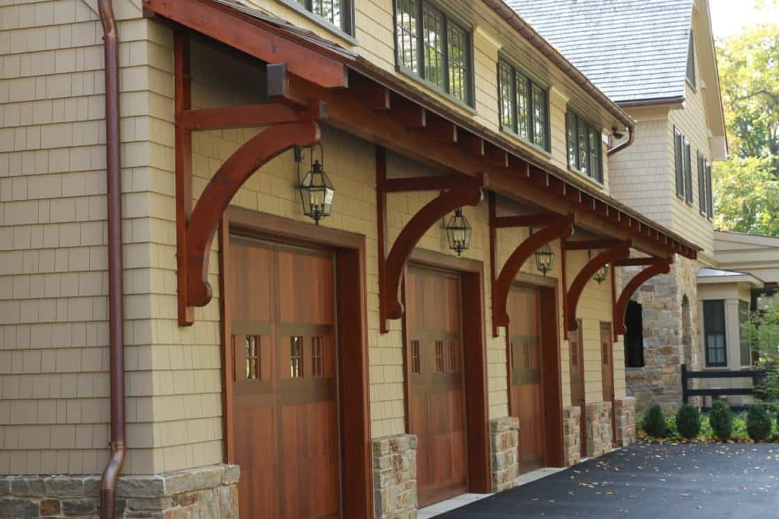 Three wooden garage doors with decorative brackets and lantern-style lights add charm to this luxury home, featuring stone accents and a blacktop driveway.