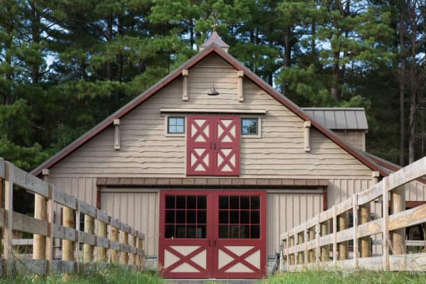 A classic bank barn with red trim and double doors, nestled between two wooden fences, stands before tall pine trees under a partly cloudy sky.