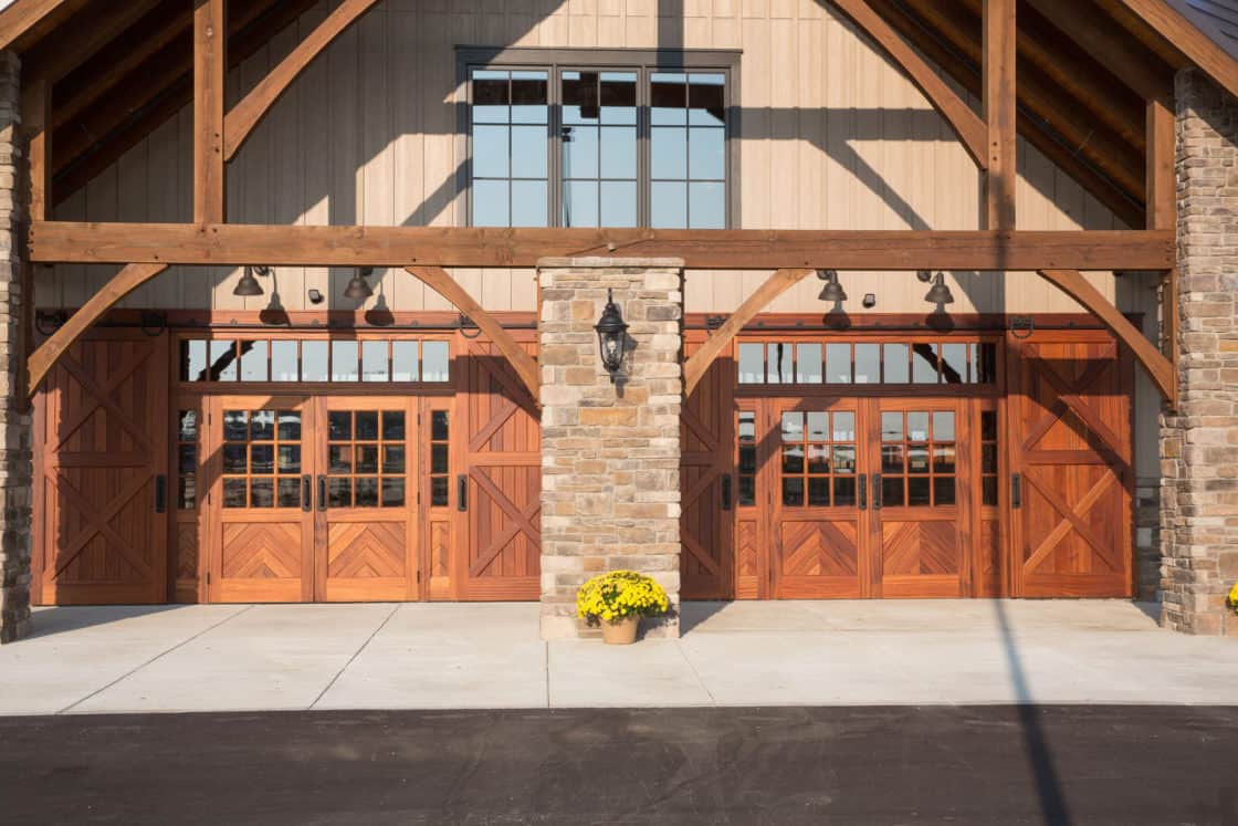 A rustic bank barn with large wooden garage doors, stone pillars, exposed wood beams, and a pot of yellow flowers by the entrance, all under bright sunlight.