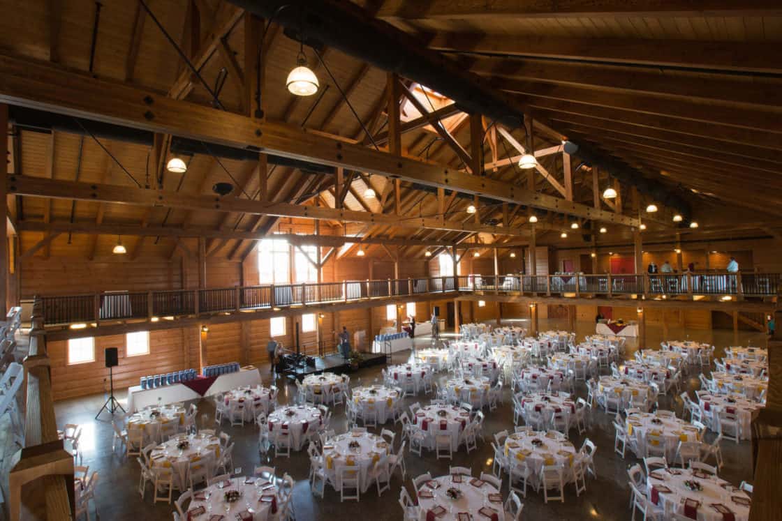 A spacious event hall with wooden beams and high ceilings, reminiscent of a luxury horse barn, filled with round tables covered in white tablecloths and surrounded by chairs. Warm lighting fills the room, perfect for a banquet or reception.