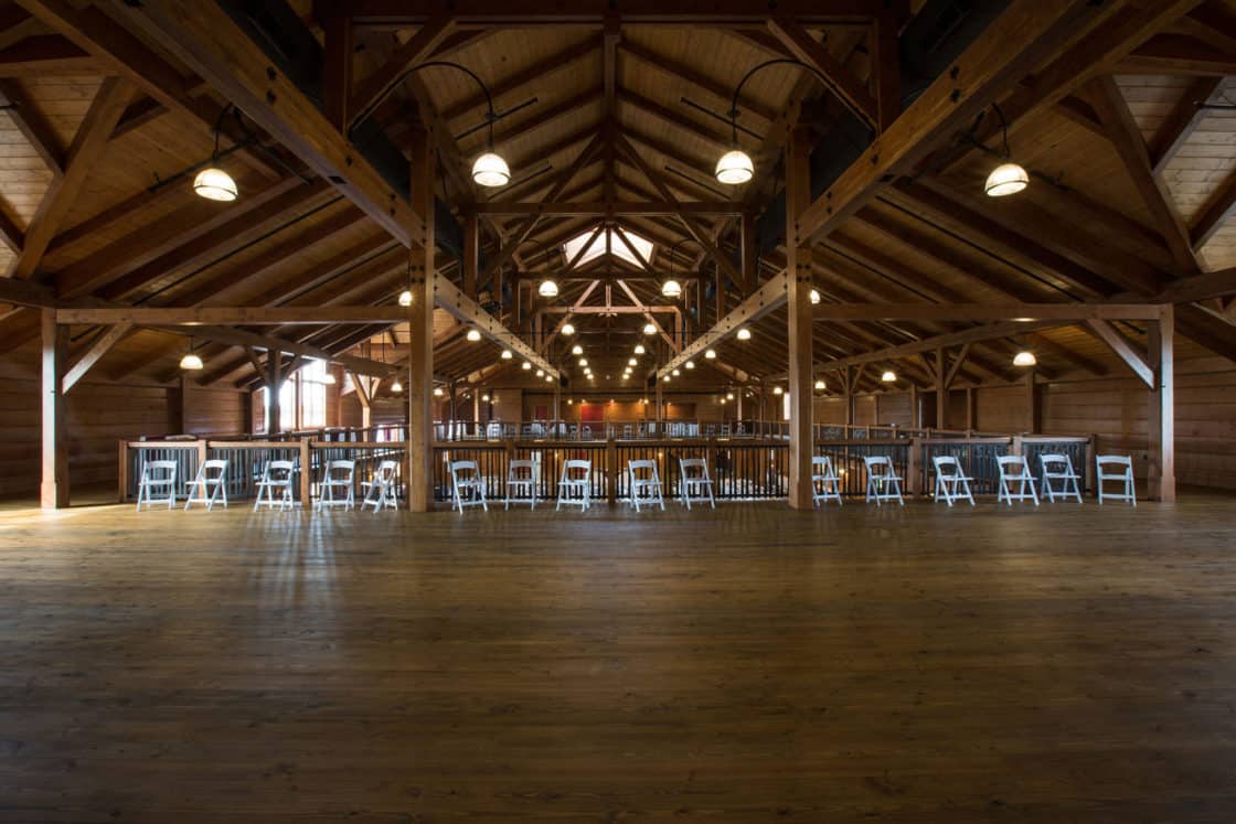 Spacious wooden party barn with exposed beams, high ceilings, and large hanging lights. Folding chairs and tables are arranged along the sides, leaving an open area in the center, with natural light streaming in.