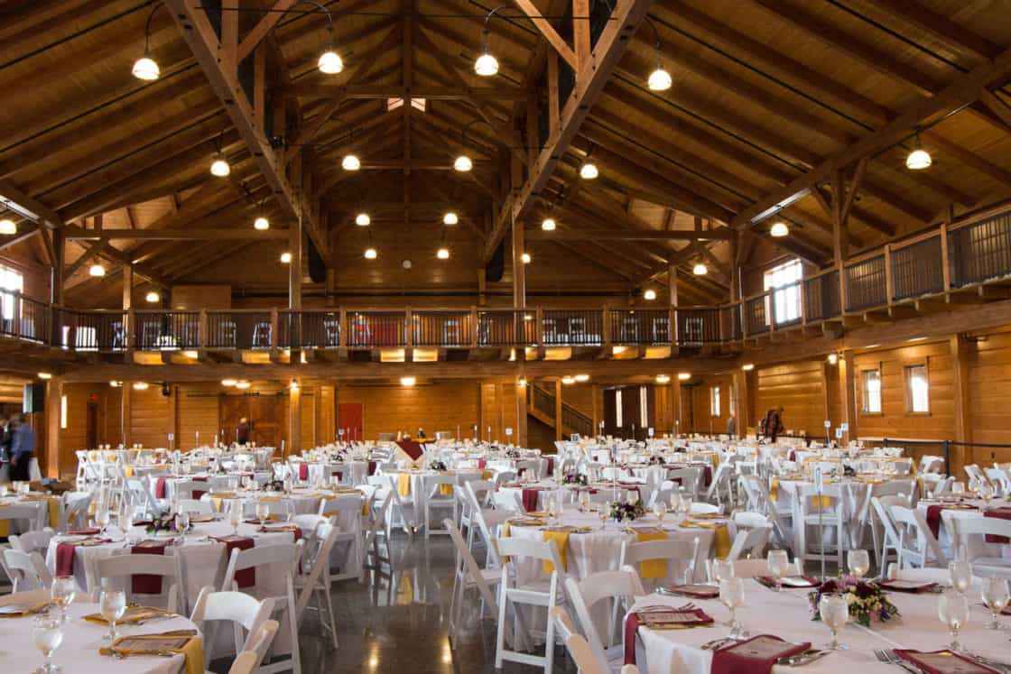 A spacious wooden hall in a renovated bank barn with a high ceiling set up for an event, featuring round tables covered with white tablecloths, red and yellow napkins, and white chairs arranged neatly.