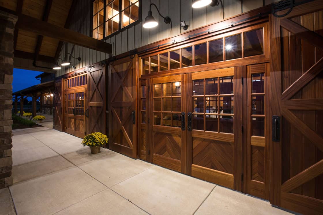 Wooden barn doors with glass window panels and black handles are illuminated by outdoor lights at dusk, evoking a luxury home vibe. Yellow potted flowers sit beside the entrance while warm light glows from inside this inviting party barn.
