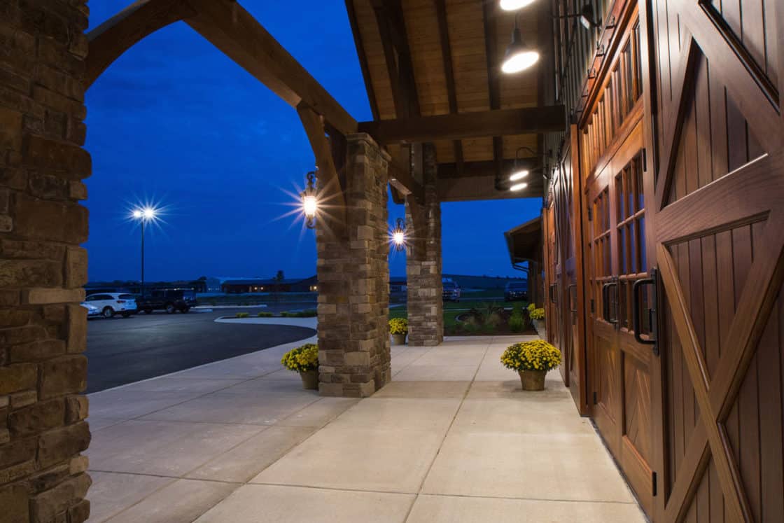 Covered outdoor walkway at dusk, featuring stone columns, wooden beams, and barn-style doors reminiscent of a luxury horse barn. Two planters with yellow flowers sit near the entrance; parking lot and streetlights visible in the background.