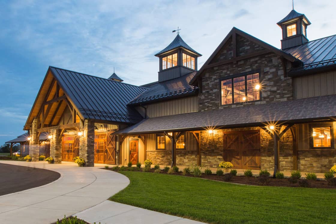 A large, rustic-style party barn with stone walls, wood beams, and warm exterior lights—complete with multiple cupolas—sits by a curved walkway and landscaped greenery at dusk.