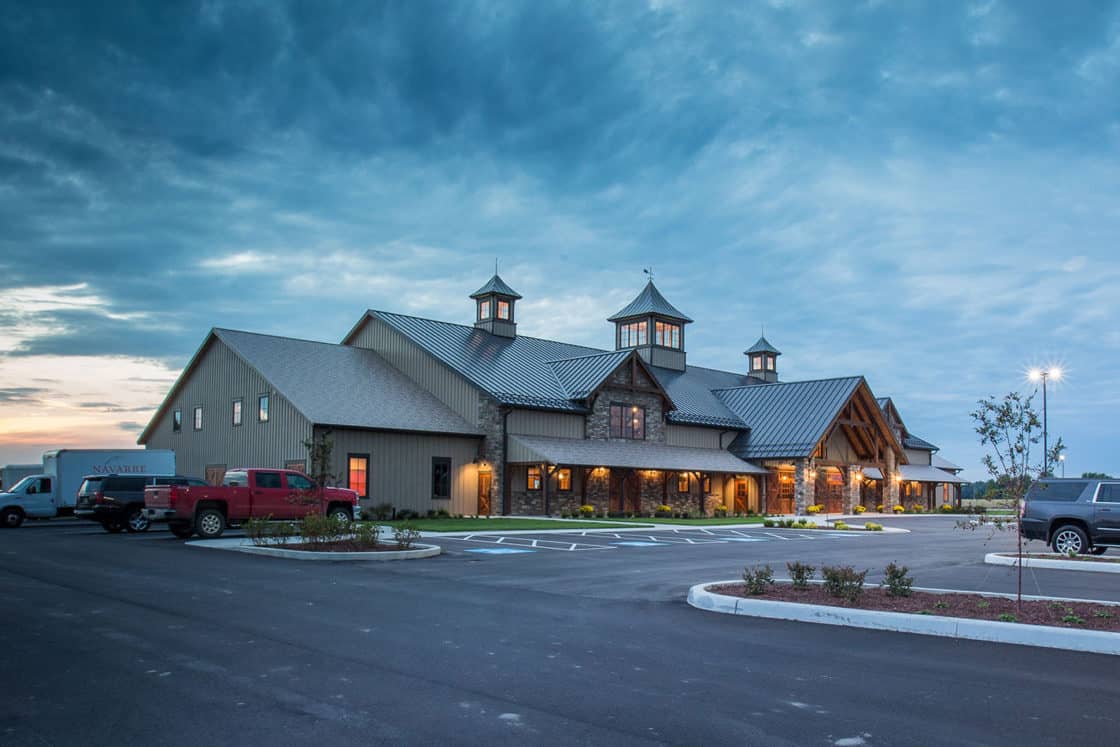 A large, modern bank barn-style building with three cupolas on the roof, warm lights glowing inside, surrounded by a parking lot with several vehicles, under a cloudy evening sky.