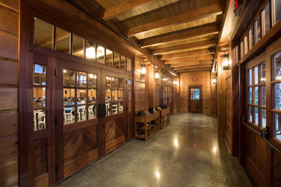 A wide wooden hallway with warm lighting, exposed beams, and glass-paneled double doors evokes the charm of a luxury horse barn. On one side, people sit at tables in a dining area visible through the windows; the polished floor gleams below.