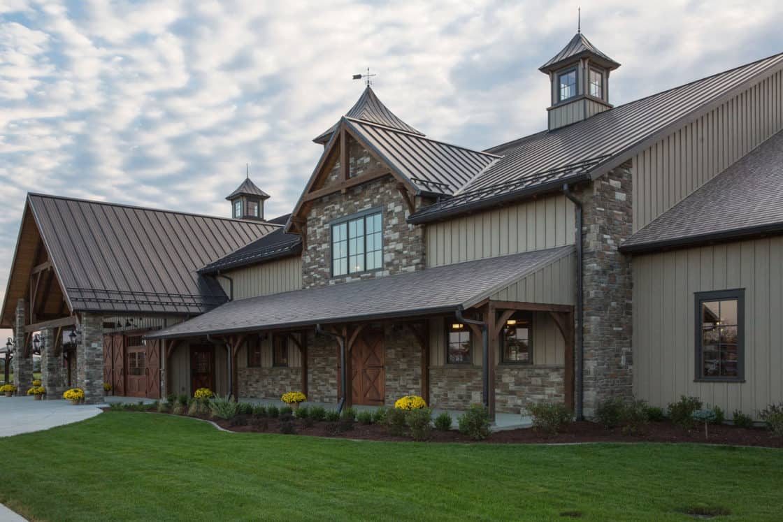 A large, rustic-style party barn with stone and wood siding, multiple peaked roofs, and cupolas, surrounded by green grass and yellow flowers under a partly cloudy sky.