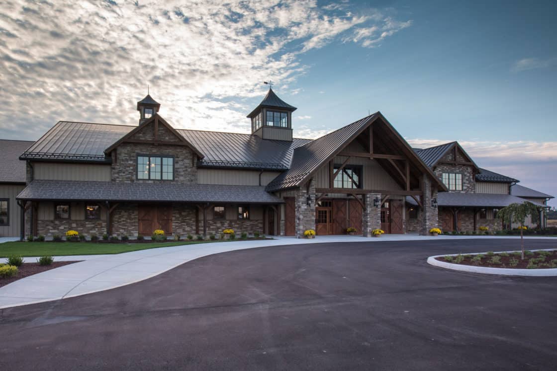A large, rustic-style luxury home with stone and wood exterior, peaked roofs, and covered entrance, set against a partly cloudy sky with landscaped grounds and a circular driveway in front.