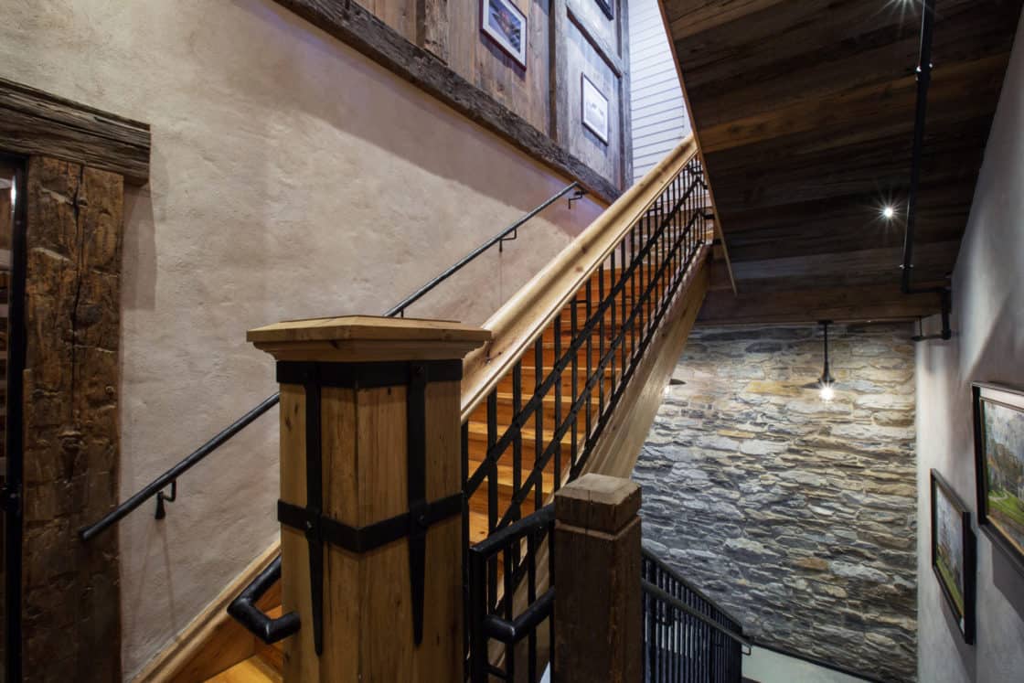 Wooden staircase with black metal railings, surrounded by rustic stone and plaster walls, wooden beams, and framed pictures on the upper wall. Soft lighting creates a warm, inviting atmosphere in this luxury home.