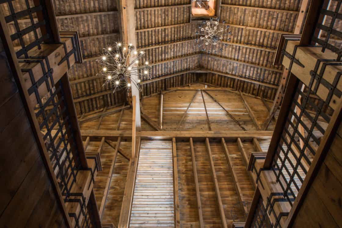 View looking up a wooden staircase inside a rustic luxury horse barn, showcasing exposed wooden beams, two modern chandeliers, and a small window at the peak of the high, vaulted ceiling.