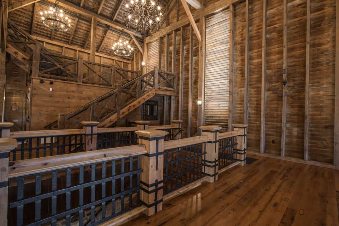 A spacious wooden interior with exposed beams, a polished wood floor, tall windows with blinds, wooden railings, and a staircase evoke the charm of a luxury home. Two ornate chandeliers hang from the ceiling.
