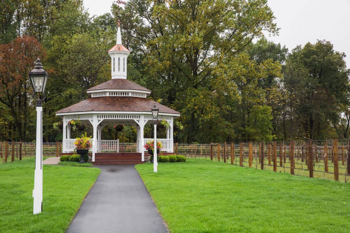 A white gazebo with a red roof sits at the end of a paved path, surrounded by green grass, wooden fences reminiscent of a luxury horse barn, and tall trees; two lamp posts stand along the walkway.