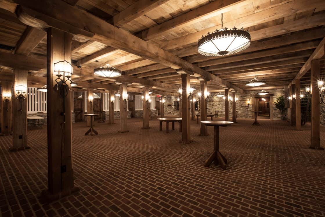 Spacious rustic party barn with brick flooring, wooden beams, stone walls, round wooden tables, and several ornate hanging light fixtures, creating a warm, inviting atmosphere.