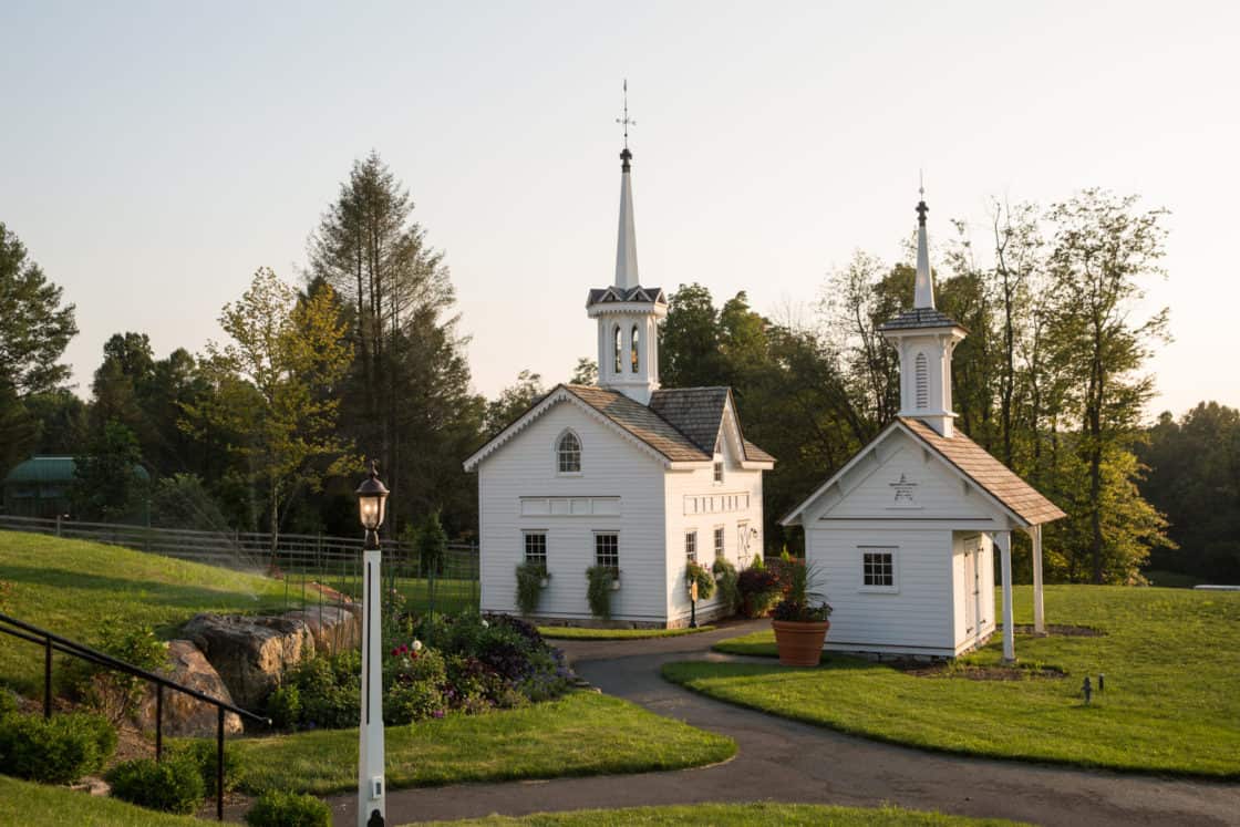 Two small white chapel-like buildings with steeples sit on a manicured lawn, surrounded by trees and garden beds under a clear sky. A paved path curves between the buildings, evoking the charm of a luxury home or rustic bank barn setting.