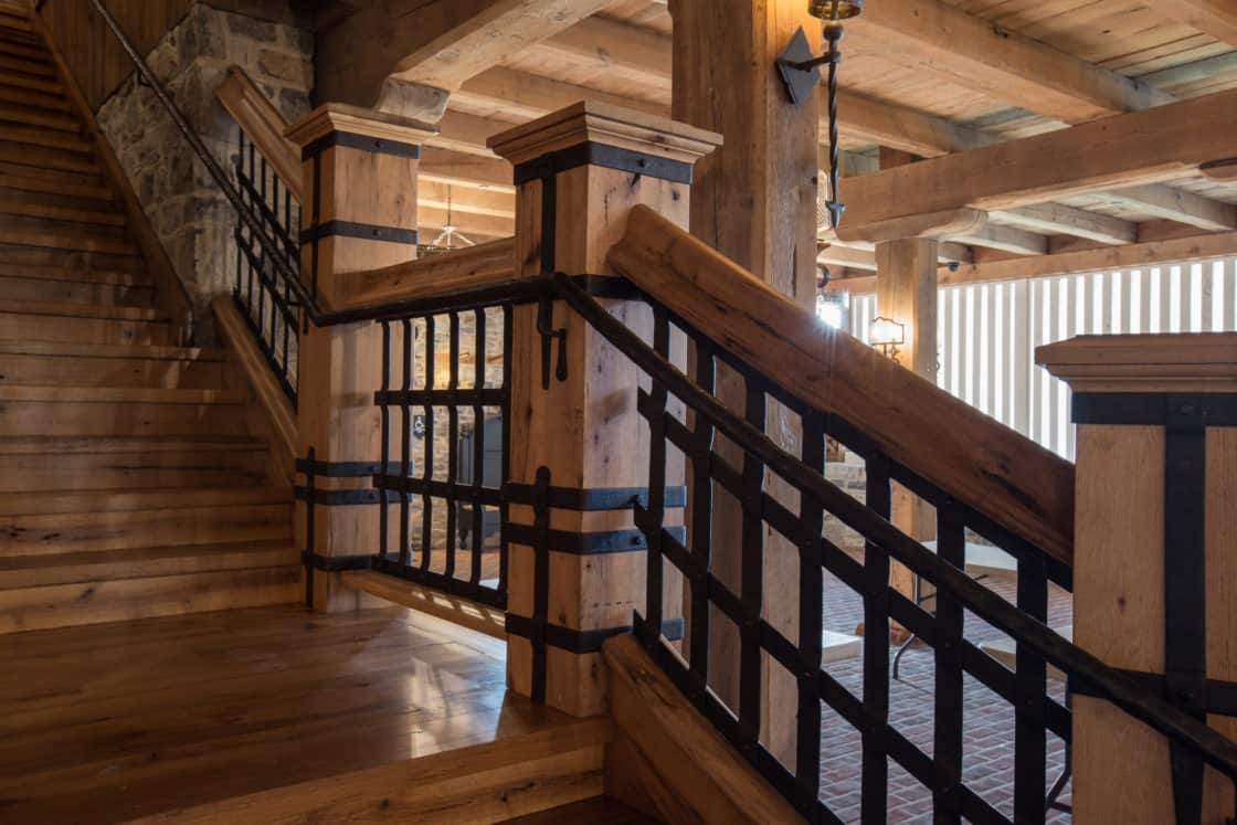 Wooden staircase with black metal railings and exposed wooden beams, creating a rustic, lodge-like interior. The area is warmly lit, highlighting the natural wood and iron details reminiscent of a luxury horse barn.