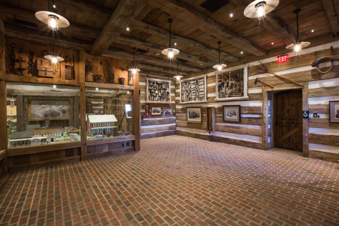 A rustic museum room with exposed wooden beams and timber walls evokes the charm of a party barn, displaying antique tools, framed photos, and dioramas under hanging lights. An open wooden door leads to another room.