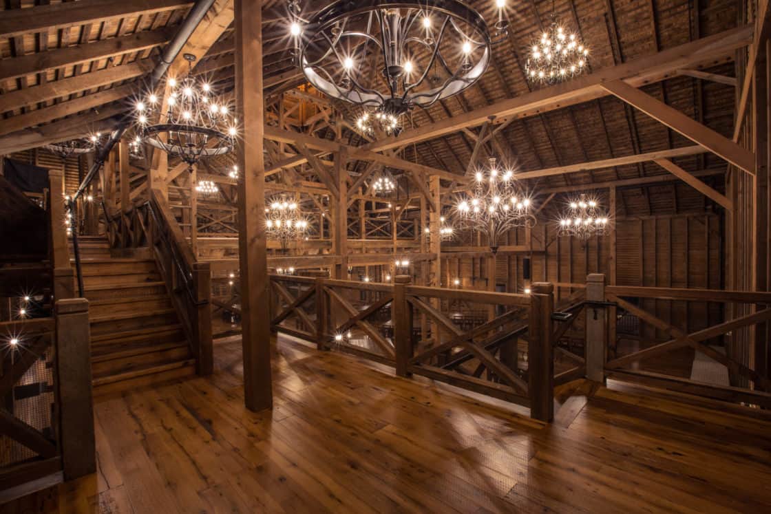 A spacious, rustic bank barn interior with wooden floors, beams, and railings, illuminated by multiple large, ornate chandeliers hanging from the high ceiling. Stairs lead to an upper loft area in this luxury horse barn setting.
