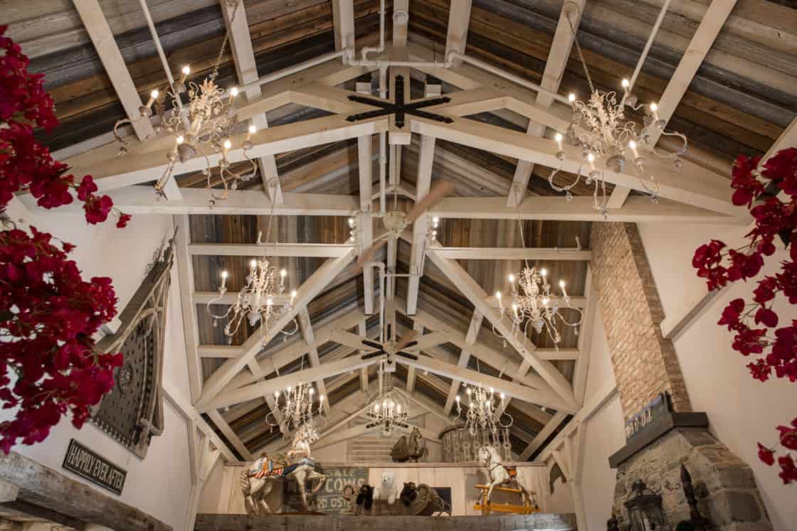 A rustic, high-ceilinged party barn with exposed wooden beams, chandeliers, red flowers on the sides, and Western-themed decor—including cowboy statues and saddles—displayed in a lofted area.