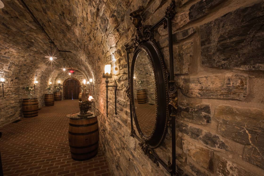 A dimly lit stone wine cellar with arched ceilings, wooden barrels lining the walls, vintage lanterns, and a large ornate mirror reflecting the rustic setting—echoing the elegance of a luxury home or party barn.