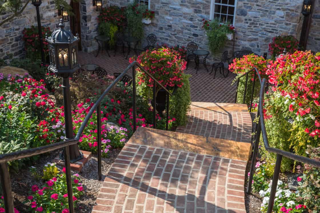 A brick staircase with black railings leads down to a patio with chairs and tables, surrounded by lush, colorful flowers and greenery beside a stone luxury home. Lantern-style lights line the pathway.