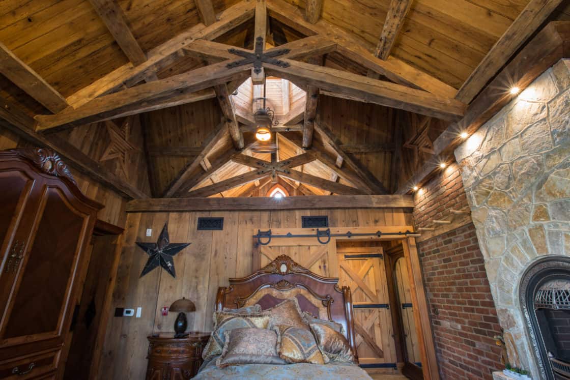 A rustic bedroom in a luxury home with exposed wooden beams, a wooden ceiling, stone fireplace, wooden doors, and an ornate bed. Warm lighting and cozy decor evoke the inviting charm of a cabin or bank barn retreat.