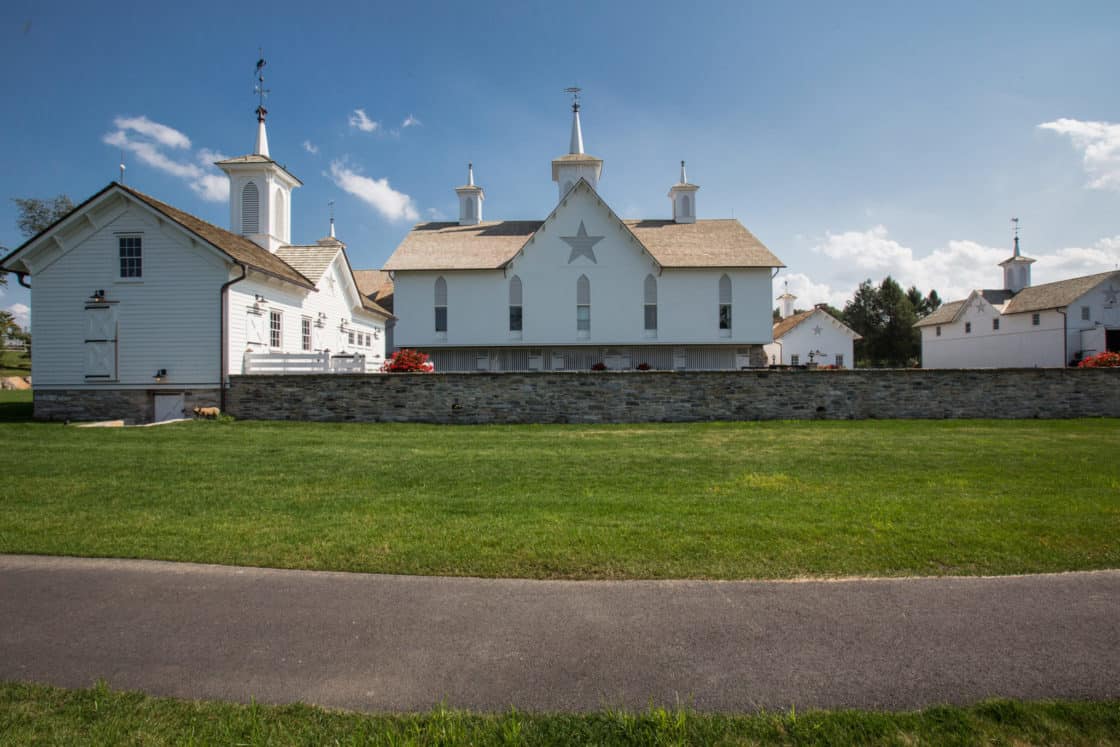 A large white bank barn with a star on the front and multiple cupolas stands behind a stone wall, surrounded by green grass and under a bright blue sky with scattered clouds.