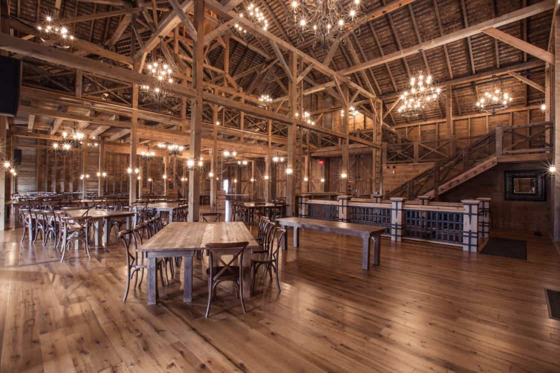 Spacious, rustic bank barn interior with wooden beams, chandeliers, long wooden tables, and chairs. Warm lighting highlights the wood floors and arched ceiling, creating an inviting atmosphere. Stairs lead to a cozy loft area.