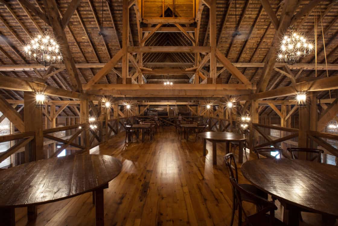 Rustic loft with exposed wooden beams, wooden floors, round wooden tables, and chairs. Warm lighting from chandeliers creates a cozy, inviting atmosphere in this spacious party barn with luxury horse barn vibes.
