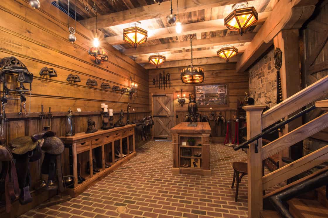 A rustic tack room in a luxury horse barn features brick floors, wooden walls and ceiling, saddles and gear on racks, a central worktable, and warm pendant lighting throughout.