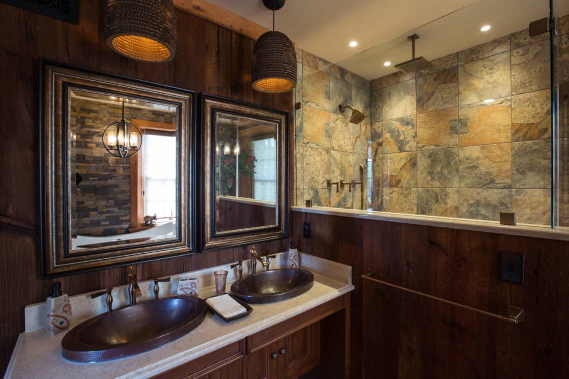 Rustic bathroom in a luxury home with double copper sinks, dark-framed mirrors, wood accents, and a tiled walk-in shower with a rain showerhead. Warm lighting creates a cozy, inviting atmosphere reminiscent of a refined bank barn.